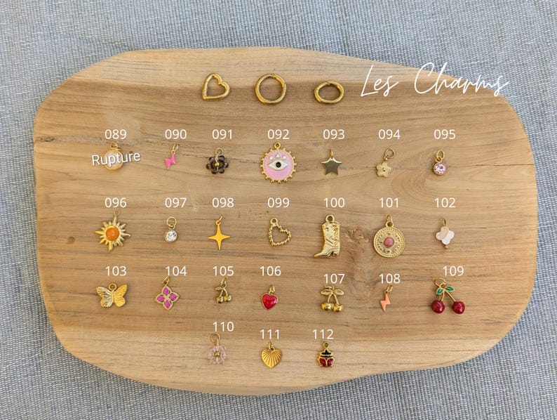 May include: A collection of gold-toned metal charms arranged on a wooden board, labeled with numbers 089 through 112. The charms include shapes like hearts, stars, flowers, a cowboy boot, an eye, cherries, and a lightning bolt, with some featuring enamel.