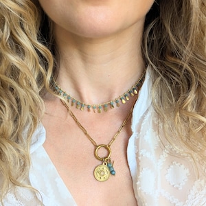 May include: A layered gold-tone necklace set consisting of a short choker featuring small blue beads and dangling gold-tone teardrop charms, and a longer chain necklace with a circular pendant containing a central stone and blue bead accents.
