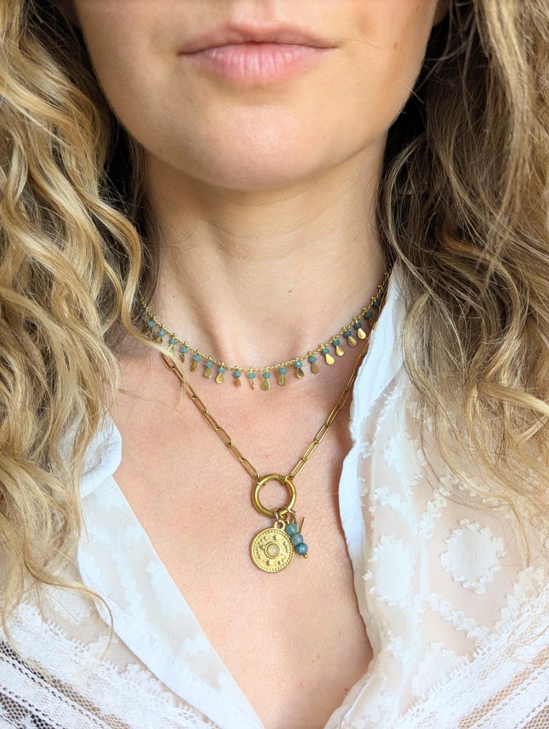 May include: A layered gold-tone necklace set consisting of a short choker featuring small blue beads and dangling gold-tone teardrop charms, and a longer chain necklace with a circular pendant containing a central stone and blue bead accents.