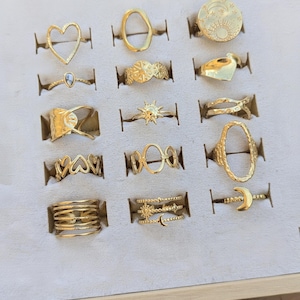 Puede incluir: Una colección de anillos dorados exhibidos en una caja de joyería. Los anillos presentan varios diseños, incluyendo corazones, soles, lunas y formas geométricas. Los anillos están hechos de un material metálico brillante.