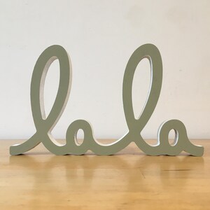 Puede incluir: Un letrero de madera verde claro con la palabra "lala" escrita en letra cursiva.