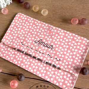 Peut inclure: Une carte rose avec des cœurs blancs imprimés dessus. La carte porte le mot "Amour" écrit en cursive noire et un bracelet perlé avec des perles brunes et blanches. La carte comporte également un petit cercle rose avec une illustration de morse et les mots "MACY JEWELS" imprimés dessus.