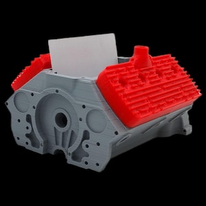 Puede incluir: Un modelo de bloque de motor de coche impreso en 3D de color gris con una tapa de válvula de plástico rojo. El bloque de motor tiene una ranura para guardar tarjetas de visita o notas.