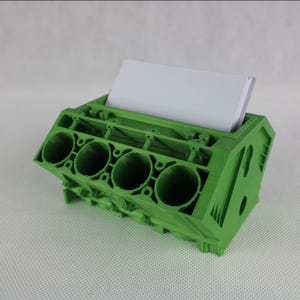 May include: Green 3D printed model of a car engine block with four cylinders. The model has a slot for holding business cards or other papers.