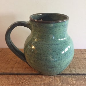 May include: A green ceramic mug with a speckled glaze and a brown rim. The mug has a rounded body and a handle.