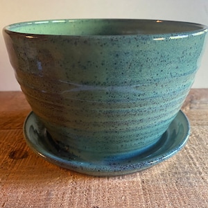 May include: A handcrafted, teal-colored ceramic planter with a matching saucer. The planter has a wide, rounded bowl shape with textured, horizontal ridges. The glaze has a speckled appearance, adding depth to the design. The saucer is round and complements the planter's aesthetic.
