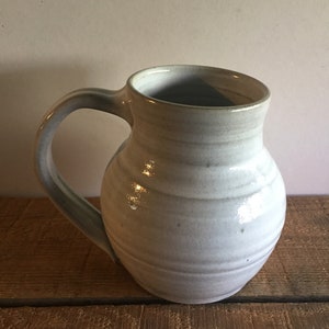 May include: A white ceramic mug with a rounded body and a handle. The mug has a slightly textured surface and a subtle glaze.