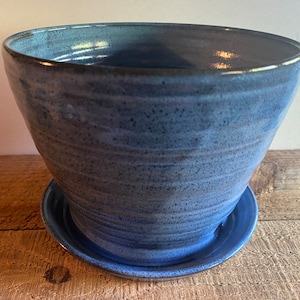 May include: A handmade, blue ceramic planter with a matching saucer. The planter has a wide, rounded shape with a textured surface. The blue glaze has variations in tone, adding depth to the piece. The saucer sits beneath the planter, providing a stable base.