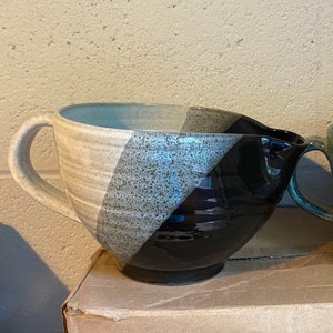 May include: A ceramic mixing bowl with a handle and spout. The bowl is divided diagonally into three sections: beige, speckled gray, and glossy black. The bowl sits on a cardboard box.