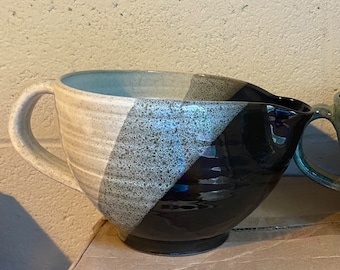 May include: A ceramic mixing bowl with a handle and spout. The bowl is divided diagonally into three sections: beige, speckled gray, and glossy black. The bowl sits on a cardboard box.