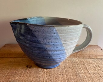 May include: A large, handmade ceramic mixing bowl with a pouring spout and handle. The bowl is divided diagonally, with a blue-gray section and a light gray section. The bowl sits on a wooden surface.