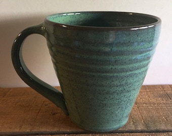 May include: A green ceramic mug with a speckled glaze and a brown rim. The mug has a wide base and a tapered shape.