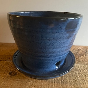 May include: A handcrafted, blue ceramic planter with a matching saucer. The planter has a wide, slightly flared shape and a drainage hole. The glaze has a speckled appearance, adding texture. The set is designed for indoor or outdoor use.