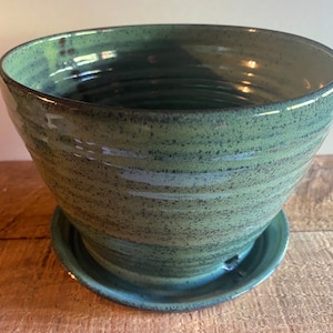 May include: A handcrafted, green ceramic planter with a matching saucer. The planter has a wide, bowl-like shape with a textured surface and a dark rim. The saucer has a small drainage hole. The planter is approximately 10 inches in diameter.