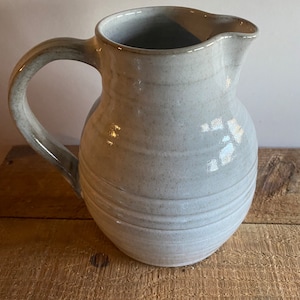 May include: A handmade, light gray ceramic pitcher with a curved handle and a pouring spout. The pitcher has a textured surface with horizontal ridges and sits on a wooden surface. The pitcher is suitable for serving water or other beverages.