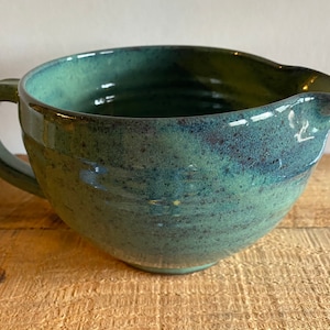 May include: A large, green ceramic mixing bowl with a speckled glaze and a handle. The bowl has a wide, rounded base and a slightly flared rim.
