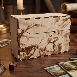 May include: A wooden card storage box with a laser-etched illustration of a fantasy character. The box is rectangular with a sliding lid and is surrounded by game cards and dice on a wooden surface.