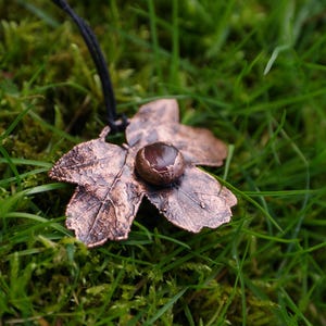 May include: A copper-colored leaf pendant with a dark brown stone center, suspended from a black cord. The pendant rests on a bed of green grass and moss, creating a natural, earthy aesthetic. The leaf design features detailed veining.