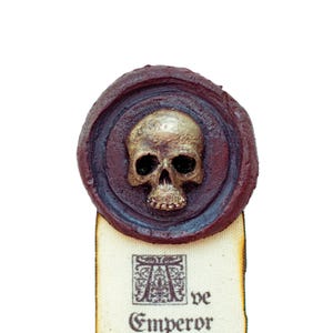 Puede incluir: Un sello de cera con un diseño de calavera en un marco circular. El marco es de color rojo oscuro con un borde dorado. La calavera es plateada con cuencas de ojos negras. El texto "Ave Emperor" está escrito en un trozo de pergamino debajo del sello.