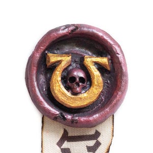 May include: A red wax seal with a gold symbol of a horseshoe with a skull inside. The seal is attached to a white ribbon with black markings.