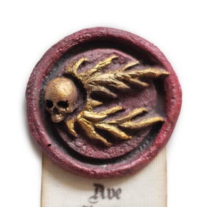 May include: A red wax seal with a gold skull and wings design. The skull has a gold crown and the wings are spread out. The seal is on a white background.