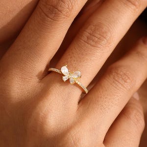 Peut inclure: Une bague dorée ornée d'un motif papillon délicat au centre, agrémentée de petites pierres claires et scintillantes. L'anneau est fin et métallique, présentant un design raffiné qui repose élégamment sur le doigt avec une finition polie.