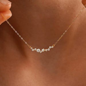 May include: A delicate gold chain necklace featuring a cluster of small, clear, round-cut stones arranged in a horizontal arc. The necklace has a fine link chain and is positioned against the neck. The text OpulenGemGB is visible at the bottom.