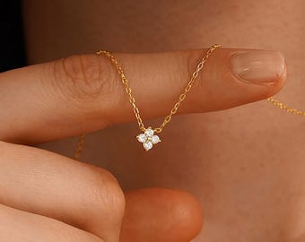 May include: A delicate gold chain necklace featuring a small, four-petaled flower pendant set with four clear, sparkling stones. The necklace rests against the skin, showcasing a thin, polished gold link chain and a dainty, symmetrical floral design.