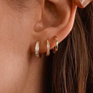 May include: Three gold hoop earrings of different designs are shown on an ear. The first hoop has a white rectangular design, the second has small clear stones, and the third is a plain gold hoop. The earrings are part of a jewelry collection.