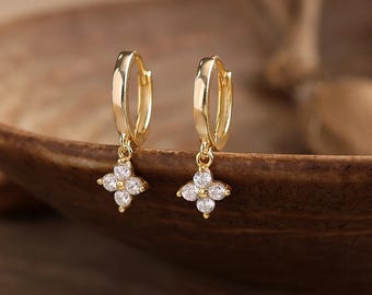 May include: A pair of gold hoop earrings featuring a small, four-petaled flower charm dangling from each hoop. Each flower charm is set with four clear, round-cut stones arranged in a cross pattern. The earrings have a polished gold finish.