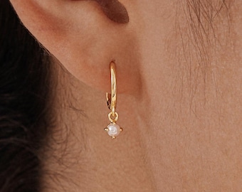 May include: A gold hoop earring with a small, white gemstone pendant. The earring is suspended from an ear. The image is a close-up, highlighting the jewellery's design and the ear's details.