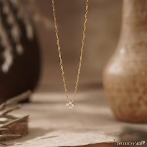 May include: A delicate gold chain necklace featuring a small, four-petaled flower-shaped pendant. The center of the pendant is set with four tiny, clear, sparkling stones arranged in a cluster, creating a subtle and refined look for this piece.