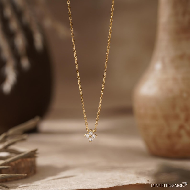 May include: A delicate gold chain necklace featuring a small, four-petaled flower-shaped pendant. The center of the pendant is set with four tiny, clear, sparkling stones arranged in a cluster, creating a subtle and refined look for this piece.