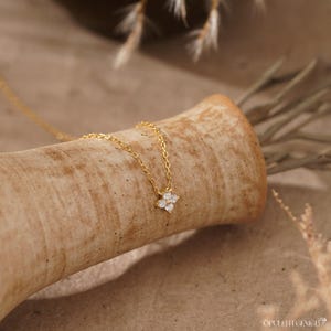 May include: A gold-toned chain necklace featuring a small, four-petaled flower pendant. The pendant is set with four clear, sparkling stones arranged in a floral pattern. The delicate metal chain connects to the center of the flower-shaped charm.
