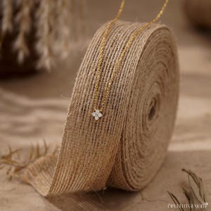 May include: A delicate gold chain necklace featuring a small, four-petaled flower pendant set with four clear, sparkling stones. The necklace is draped over a roll of textured, natural-colored burlap ribbon, highlighting the fine metal link chain.