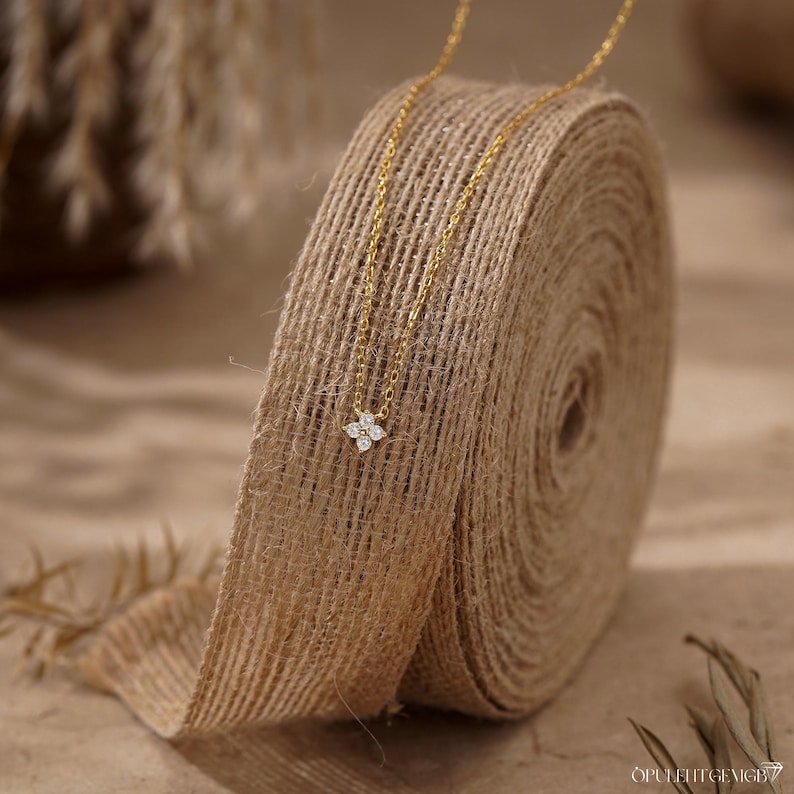 May include: A delicate gold chain necklace featuring a small, four-petaled flower pendant set with four clear, sparkling stones. The necklace is draped over a roll of textured, natural-colored burlap ribbon, highlighting the fine metal link chain.