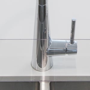 May include: A clear acrylic splash guard with two suction cups on each side, designed to protect a kitchen sink from splashes. The guard is installed above a stainless steel sink with a chrome faucet.