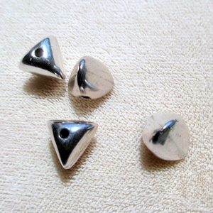 May include: Four small, smooth, silver-colored metal beads shaped like rounded triangles. Three of the beads feature a visible circular hole for threading, while the fourth bead is positioned to show its solid, rounded, and reflective surface.