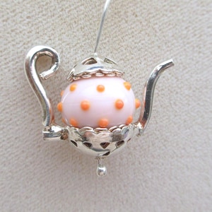 May include: A silver-toned teapot-shaped pendant featuring a light pink spherical body decorated with small orange polka dots. The metal frame includes a curved handle, a slender spout, and decorative filigree detailing on the lid and base.