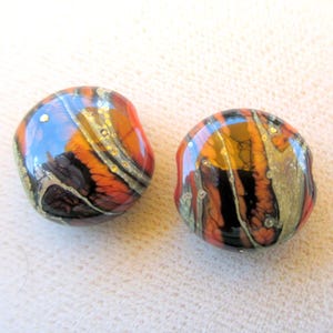May include: Two round glass beads featuring a marbled pattern of orange, black, and metallic gold swirls. The surface of each bead is smooth and glossy, displaying a complex, layered design with distinct color transitions and small gold flecks.