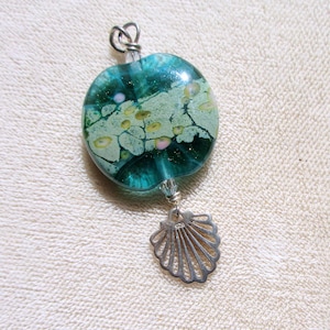 May include: A handcrafted pendant featuring a teal and green glass bead with a silver seashell charm. The glass bead has a unique pattern with yellow and pink accents. The pendant is suspended from a silver bail.