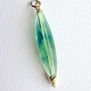 May include: A pendant with a teardrop-shaped, translucent green and blue stone. The stone is capped with gold-colored metal at both ends, with a loop at the top for hanging. The pendant is set against a white background.