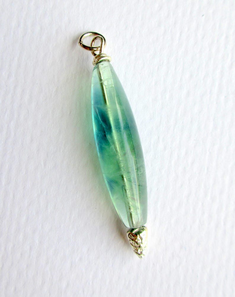 May include: A pendant with a teardrop-shaped, translucent green and blue stone. The stone is capped with gold-colored metal at both ends, with a loop at the top for hanging. The pendant is set against a white background.