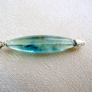 May include: A translucent, elongated pendant with a silver bail and end cap. The pendant is a mix of blue and green hues, with a polished, smooth surface. The jewelry piece is likely a necklace pendant.