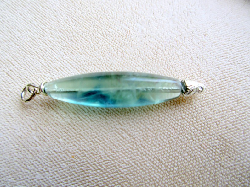 May include: A translucent, elongated pendant with a silver bail and end cap. The pendant is a mix of blue and green hues, with a polished, smooth surface. The jewelry piece is likely a necklace pendant.