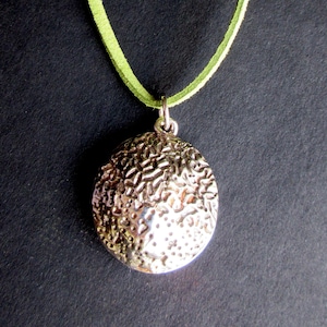 May include: A circular silver-toned pendant with a highly textured, cratered surface suspended from a bright lime green cord. The metal pendant features an irregular, pitted finish that reflects light across its uneven, rounded metallic face.