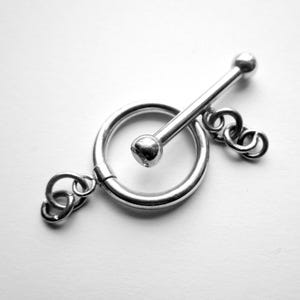 May include: A silver-toned toggle clasp, featuring a circular ring with a bar and ball closure. The clasp is connected to a small chain of interlocking rings. The image is in black and white.