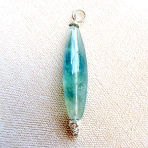 May include: A pendant featuring a long, teardrop-shaped fluorite stone with shades of teal and green. The stone is capped with a silver-colored metal setting and a small loop for a chain.
