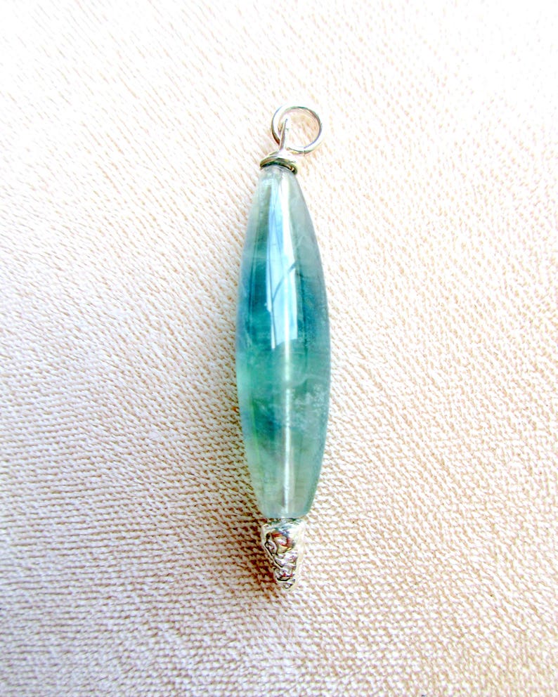 May include: A pendant featuring a long, teardrop-shaped fluorite stone with shades of teal and green. The stone is capped with a silver-colored metal setting and a small loop for a chain.