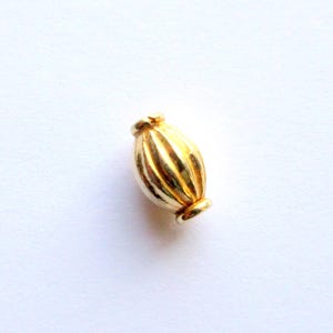 May include: A gold-colored, ribbed bead with a rounded, elongated shape. The bead has a small, circular detail at each end. The bead is likely used for jewelry making or other craft projects.